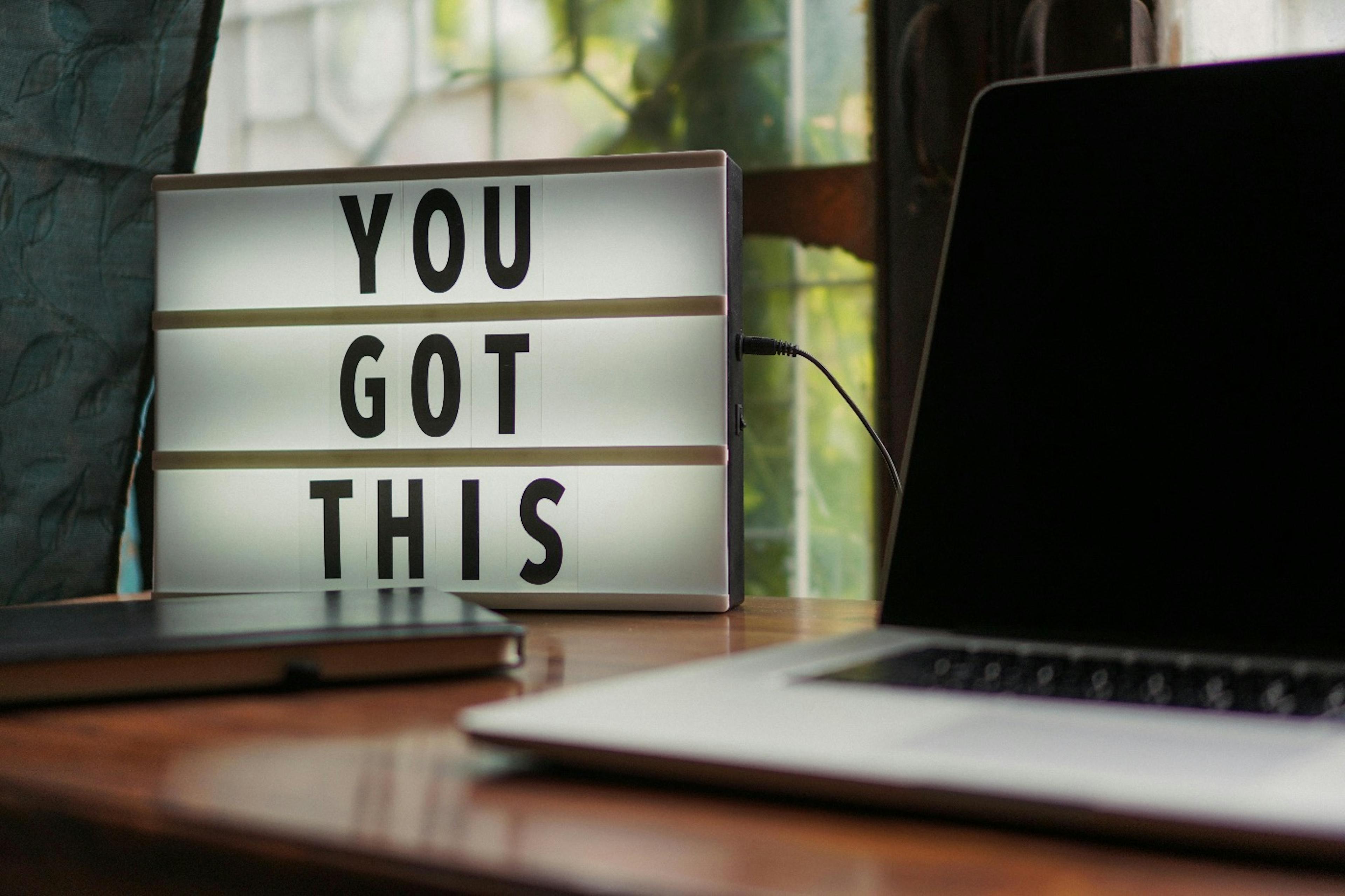 A laptop, notebook and a sign on the table. Sign says "You got this".