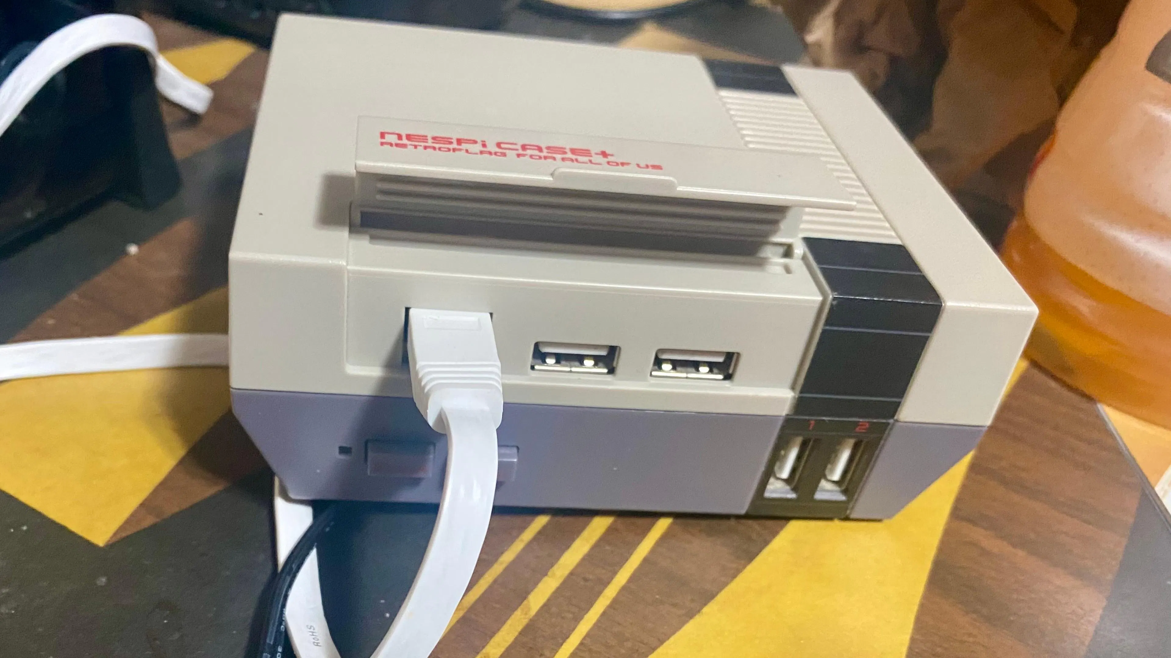 A small replica of a Nintendo Entertainment System (NES) case, with "NESPi Case" on an upturned cartridge hinge, with a white ethernet cable plugged into the front, on a wooden table with yellow and black chess-themed shapes.