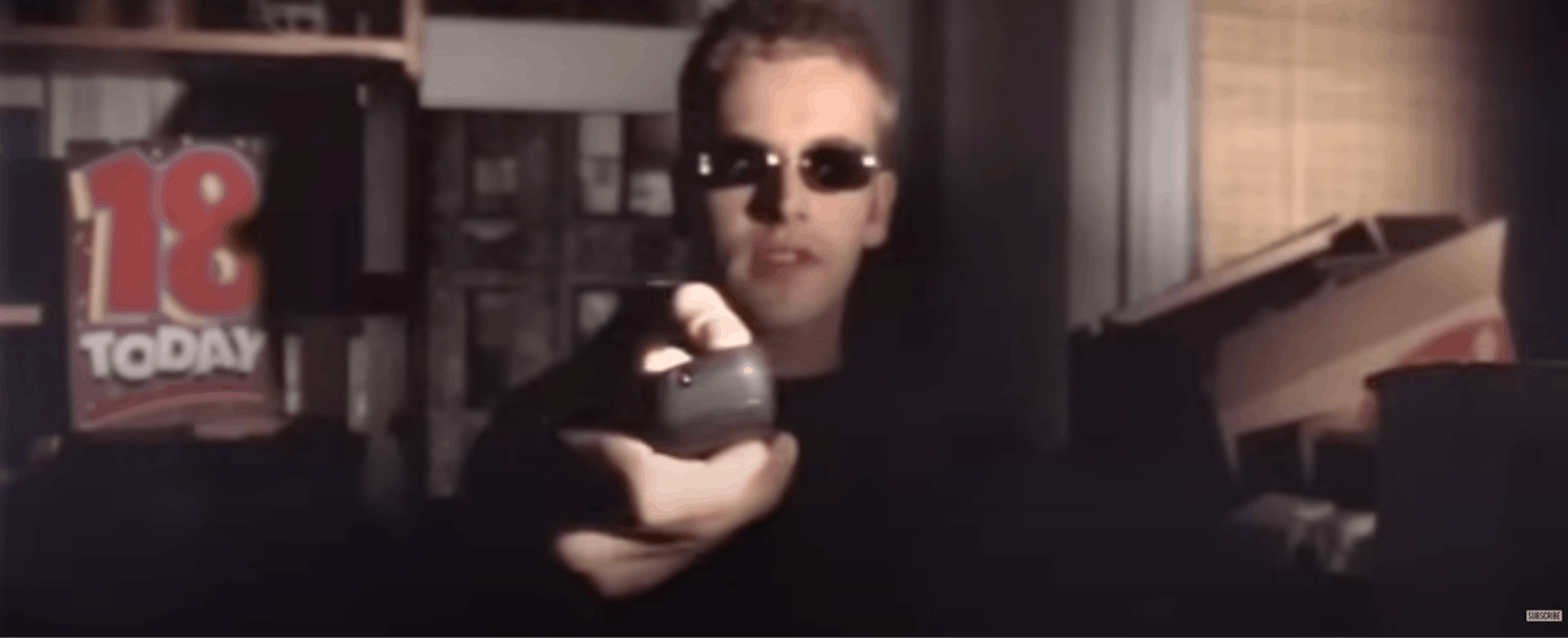 Scene depicting a character holding a remote control, wearing sunglasses in a dimly lit room. Hackers (1995). MGM Studios. Image source: YouTube – Rotten Tomatoes Classic Trailers.