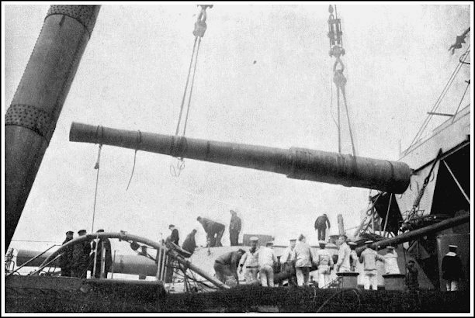 A 12-inch gun being lowered into its place in the turret of a warship by a gigantic sheer-leg crane, one leg of which is partly visible on the left of the picture.